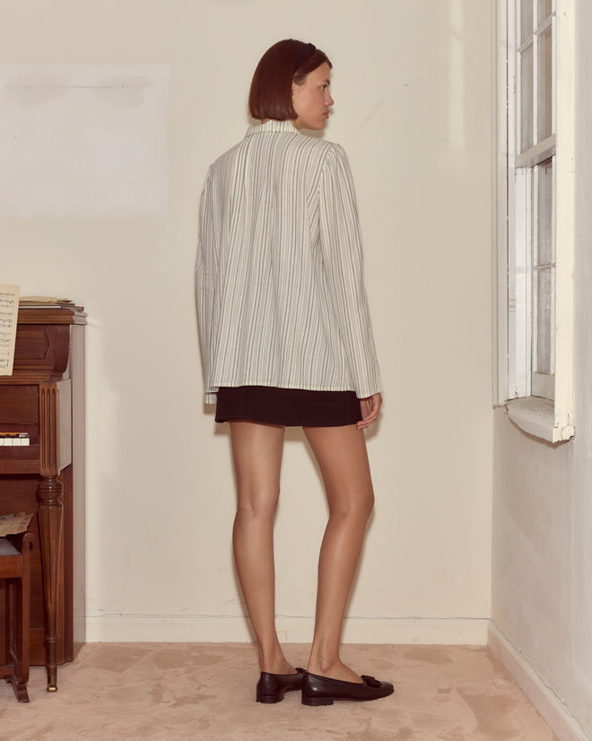 A woman in the Electric Light Blouse, featuring ivory frog closures and stripes, pairs it with a black skirt while standing in a beige room near a window and an upright piano.