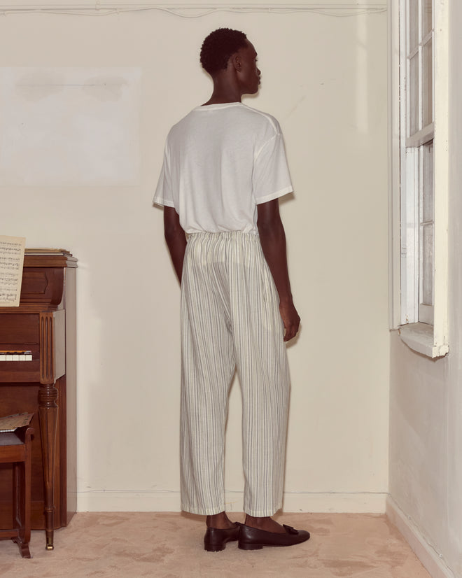 A person stands indoors near a piano and window, facing away and wearing a white t-shirt with Electric Light Pajama Pants featuring light stripes and a drawstring waist.