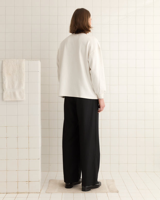 Someone wearing an Embellished Lion Crewneck and black pants stands on a tiled floor, facing away, with rhinestones subtly glimmering in the light.