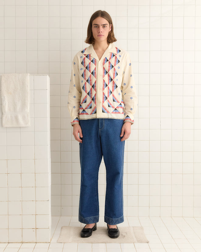 A person in an Embroidered Peak Long Sleeve Shirt and blue jeans stands in a tiled room beside a towel on a holder.