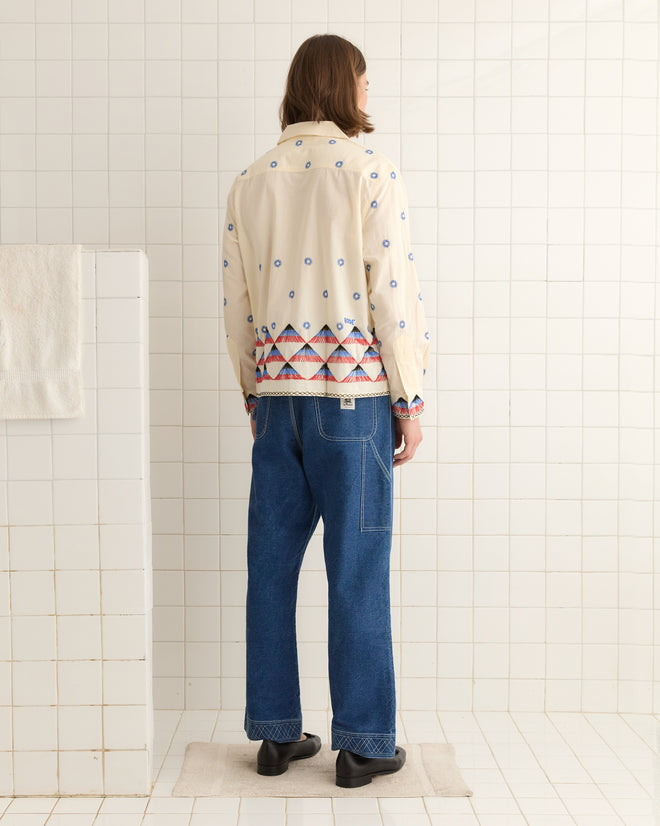 Wearing an Embroidered Peak Long Sleeve Shirt and blue jeans, a person stands on a small rug facing a tiled bathroom wall.