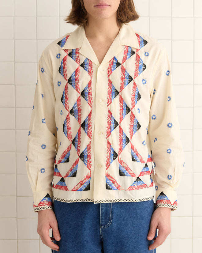 A person wearing an Embroidered Peak Long Sleeve Shirt with 1950s geometric and floral designs stands in front of a tiled wall.