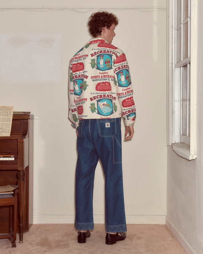 A man stands facing a corner, wearing blue jeans and the Enrichment Jacket, next to a piano and a window.