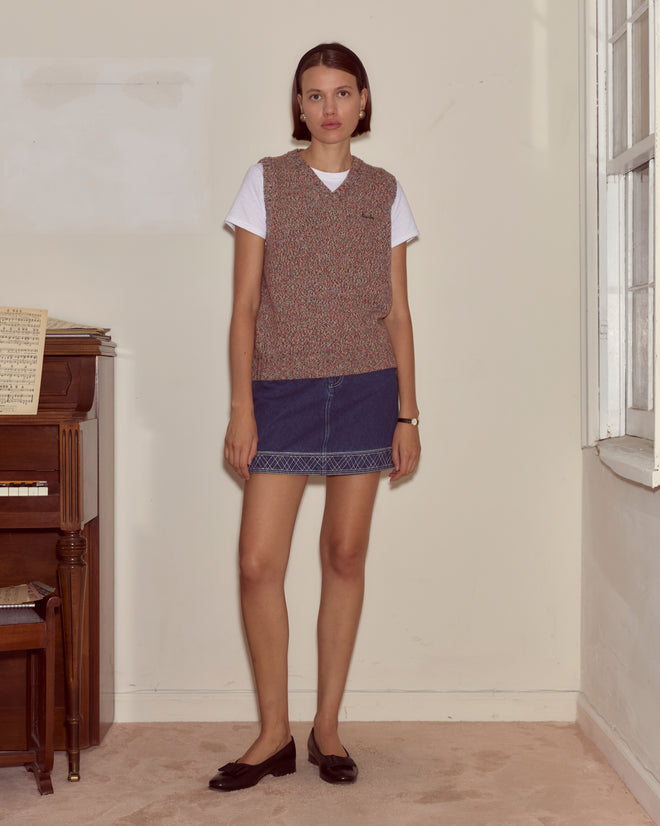 A woman stands indoors by a piano, wearing the Falls Vest - Multi from the latest women's knitwear collection, styled with a white shirt, denim skirt, and loafers.