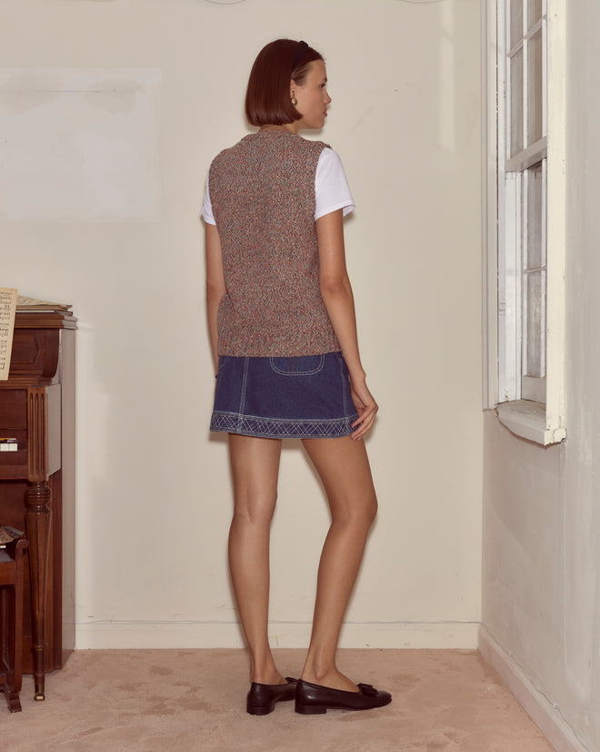 A woman stands indoors by a window, wearing the Falls Vest - Multi—a luxe wool cashmere V-neck sweater vest—over a white shirt, paired with a denim skirt and brown loafers.