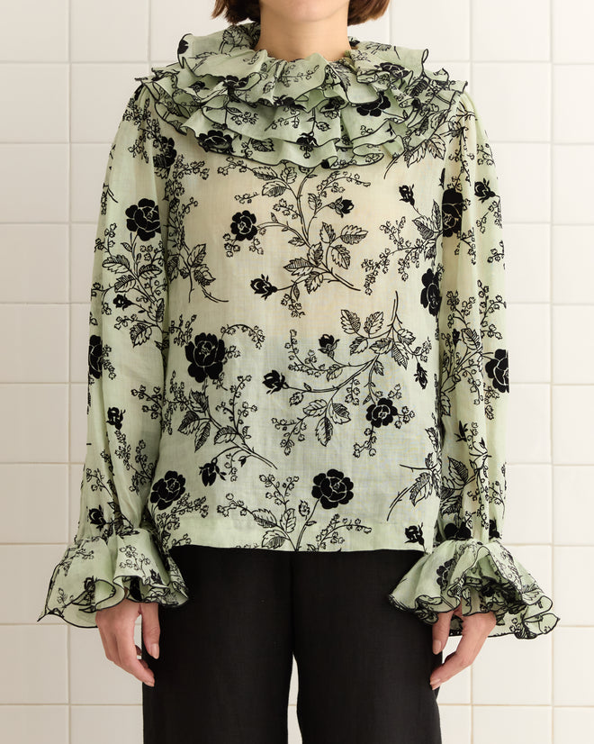 The person stands against a tiled wall, wearing the Flocked Sweetunia Top—a light green floral 1970s blouse with tiered ruffles, featuring a ruffled collar and cuffs.