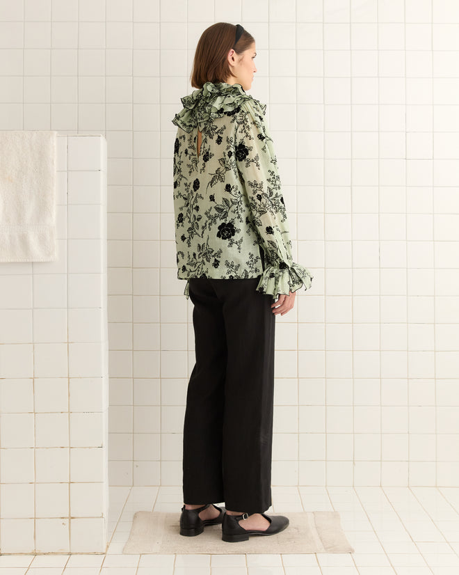 A person in the Flocked Sweetunia Top with tiered ruffles and black pants stands gracefully in a tiled room.