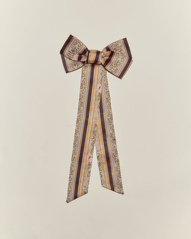 The Floret Brocade Bow Barrette features long, patterned ribbons in gold, brown, and pink, creating a decorative handmade accessory on a plain background.