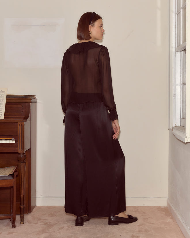 A woman in the Flounce Blouse - Black stands by a piano, facing away from the camera.