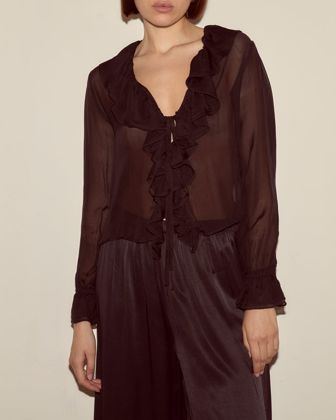 Woman wearing the Flounce Blouse - Black, a sheer silk long-sleeve ruffled French blouse, paired with matching wide-leg pants, standing against a plain wall.