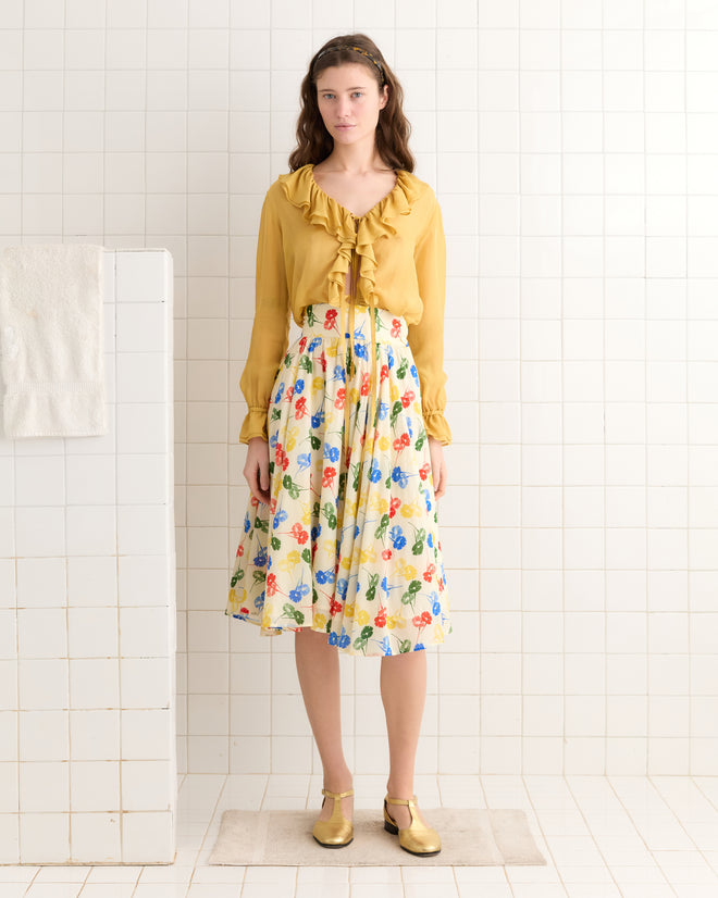 A woman in a ruffled yellow blouse and the Flower Stamp Skirt stands on a tiled floor in a white-tiled room, showcasing her timeless style.
