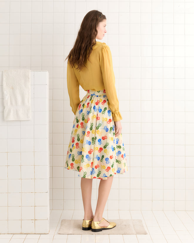 A woman in a yellow blouse and the Flower Stamp Skirt, made of 100% cotton with a floral design, stands in the white tiled bathroom, facing away.