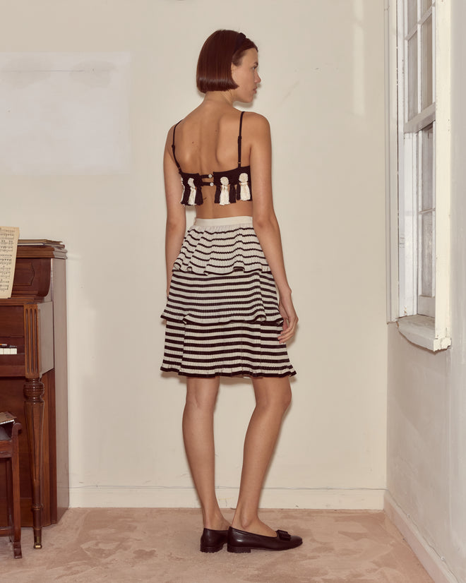 A woman in a Folly Bra and a striped skirt stands sideways near a piano and a window in a simple room.