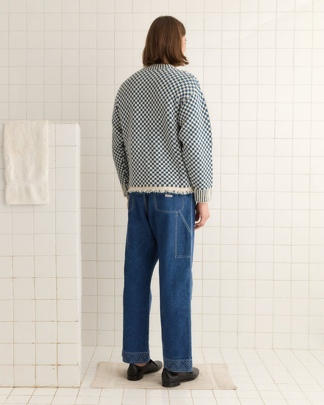 Someone with long hair in a blue Fringe Check Pullover and jeans stands on a tiled floor, facing away.