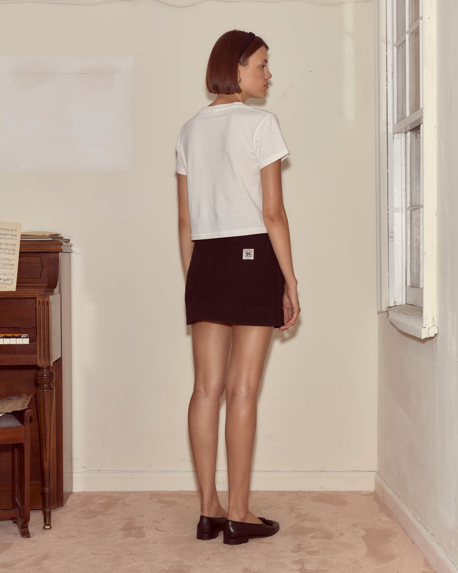 A woman in a Geneva Tee and black skirt stands by a window in a minimal room, evoking the look of a vintage photo album.