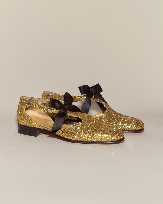 Glitter Theater Shoes - Gold: gold glittery Mary Jane shoes made from glitter leather, featuring black ribbon bows, displayed on a plain light background.