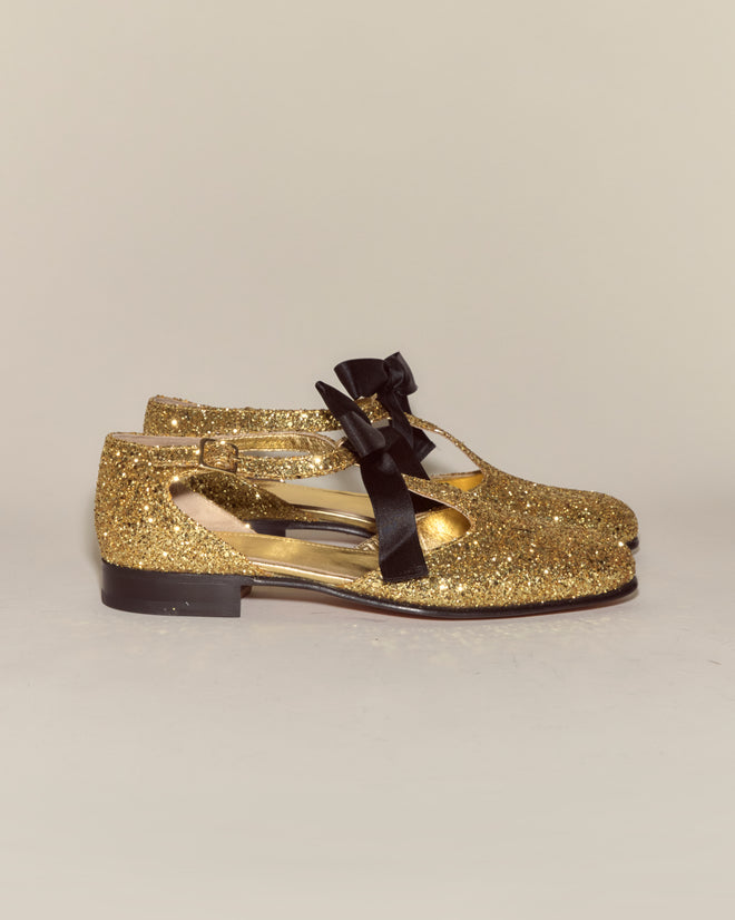 The Glitter Theater Shoes - Gold are Mary Jane-style leather shoes with gold glitter and black ribbon bows, inspired by classic midcentury dancing shoes, shown on a plain background.