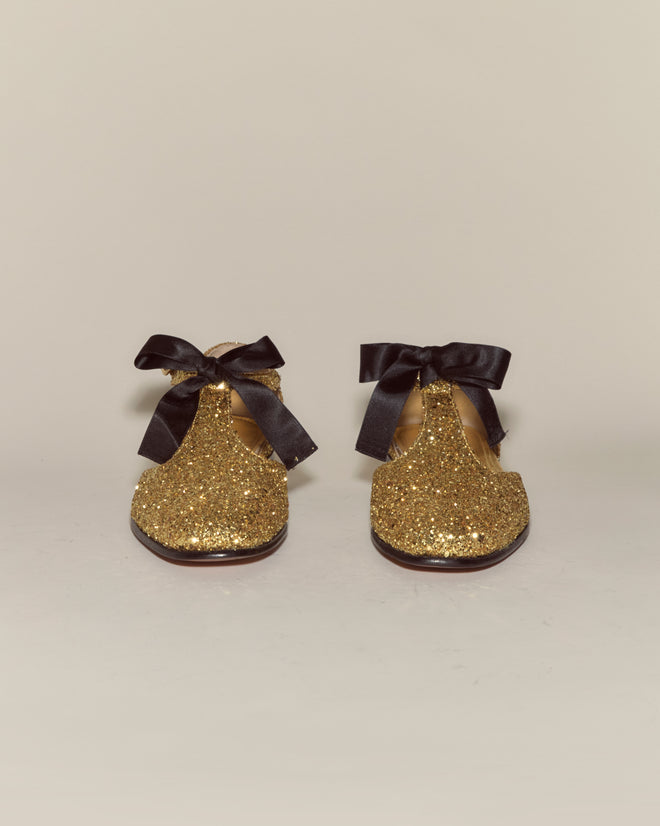 The Glitter Theater Shoes - Gold feature gold glitter leather and black ribbon bows, designed in a style reminiscent of midcentury dancing shoes, shown from the front against a plain background.