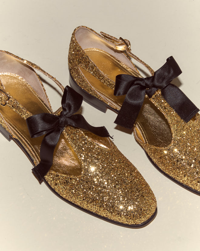 Glitter Theater Shoes - Gold: gold glittery T-strap shoes with black bows and ankle straps, inspired by midcentury dancing shoes, shown on a light background.