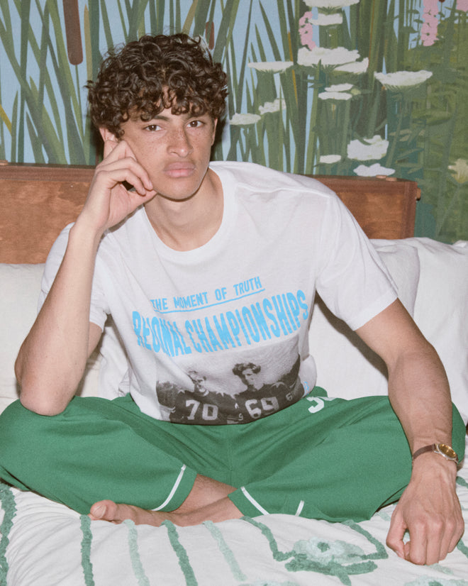 A young man with curly hair sits cross-legged on a bed, wearing green Sophomore Sweatpants and a heavyweight graphic T-shirt.