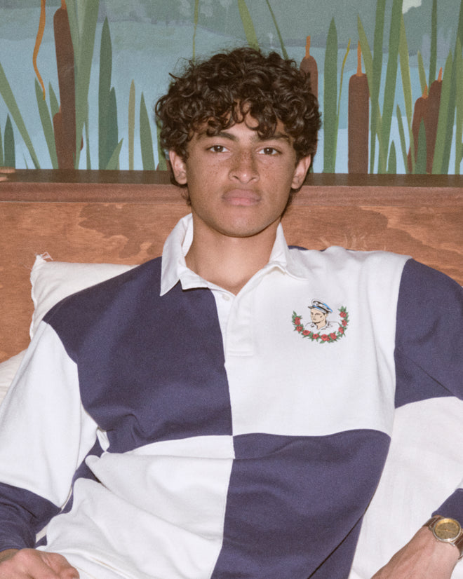 A young man with curly hair sits on a bed wearing the Grandfather Polo, a classic blue and white 1980s-inspired rugby shirt with a crest, made from 100% cotton.