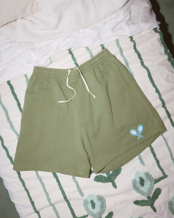 Beaded Racquet Patch Shorts in olive green with a drawstring, blue tennis racket logo, and novelty tennis patches, displayed on a patterned bedspread.