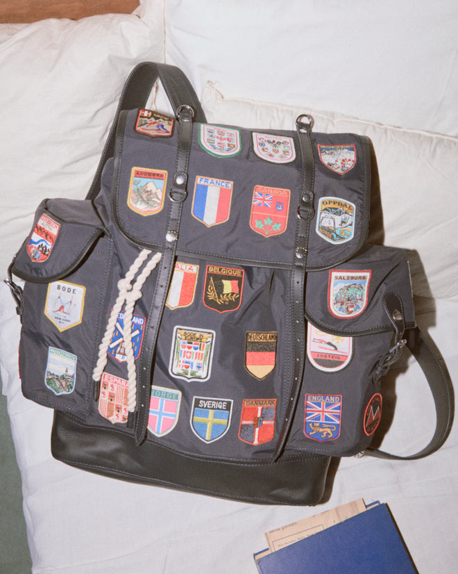 The Interrail Rucksack, a vintage-inspired black backpack adorned with colorful country patches, rests on a white cushion.