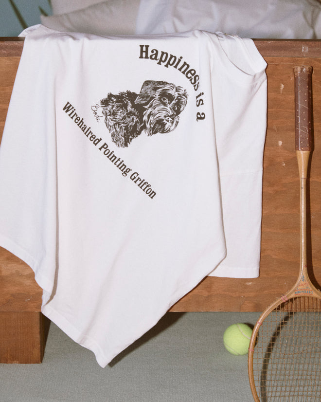 The Happiness Tee, a white 100% cotton shirt featuring a cute dog graphic and “Happiness is a Wirehaired Pointing Griffon,” rests on a wooden bench.