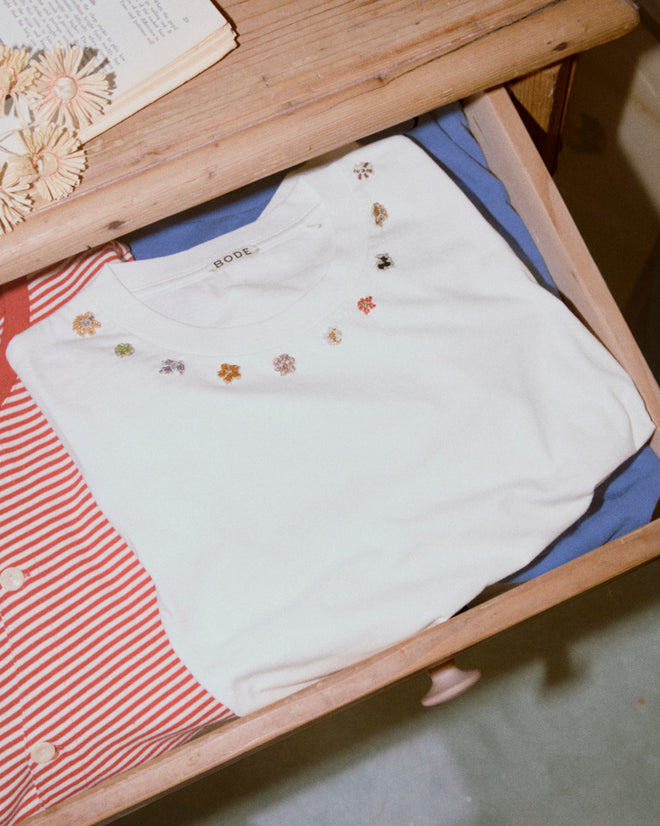 The Beaded Floral Necklace Tee, a white 100% cotton T-shirt with small embroidered flowers, is folded in a wooden drawer next to other colorful clothes.