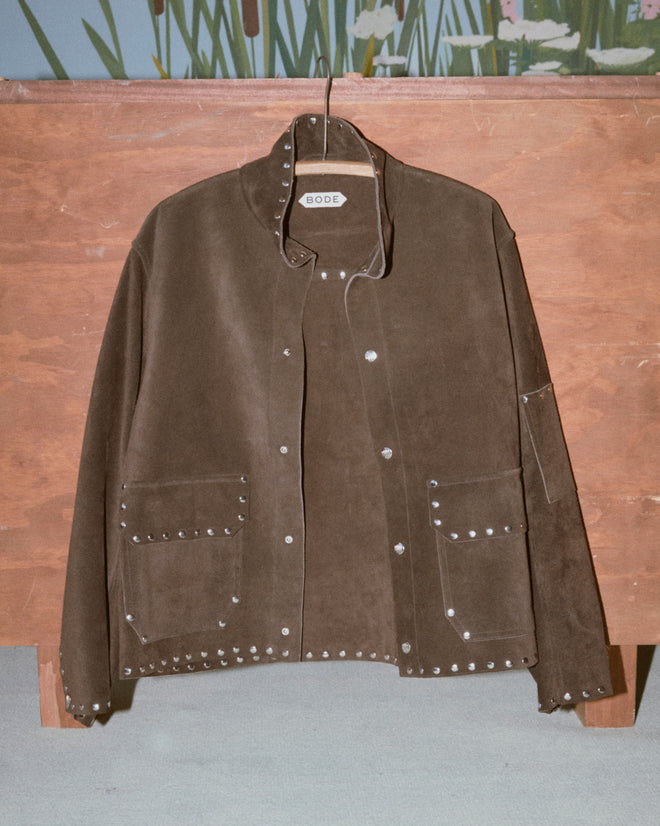 The Studded Welding Jacket - Brown features suede material and silver studs, evoking a metal shop vibe. Displayed on a wooden backdrop with the BODE label visible.