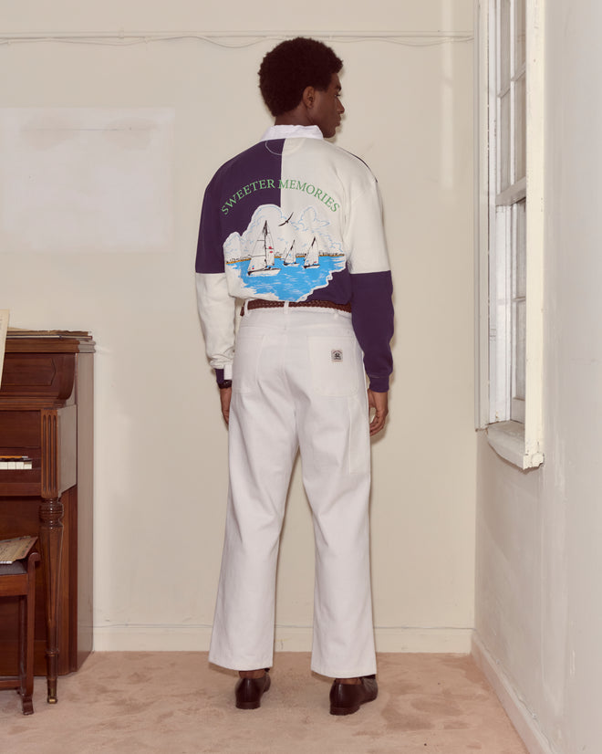 A person stands indoors facing a wall, wearing white pants and the Grandfather Polo, a cotton sweater with a sailboat graphic and text for a classic 1980s vibe.