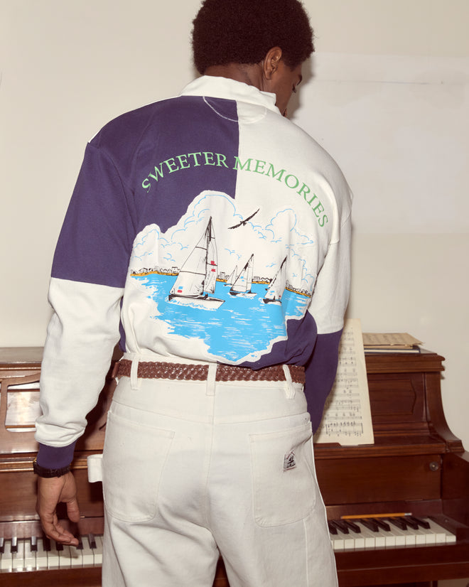 A person wearing the Grandfather Polo and white cotton pants stands by a piano with sheet music, their back to the camera.
