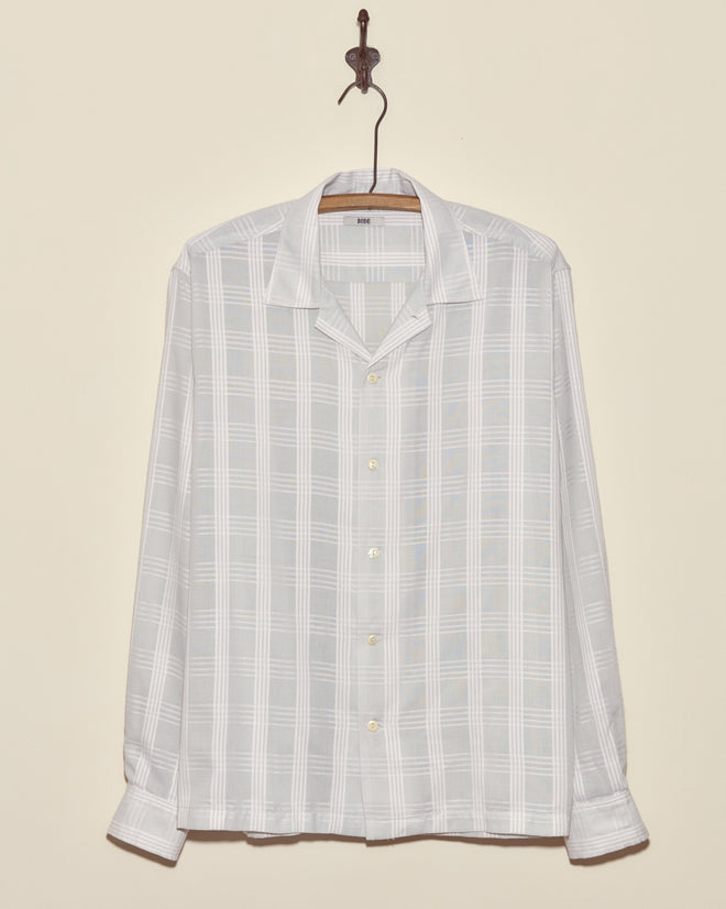 The Grayscale Long Sleeve Shirt - L/XL, made from deadstock plaid shirting fabric, hangs on a wooden hanger against a beige wall.