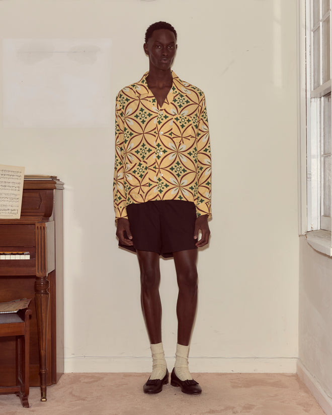 A person stands indoors beside a piano, wearing the Greer Long Sleeve Shirt with a geometric pattern, paired with black shorts, socks, and loafers.