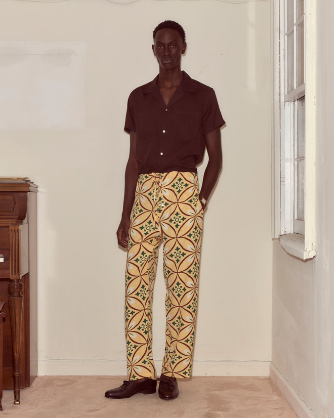A man stands indoors wearing a dark short-sleeve shirt, Greer Pajama Pants with a botanical print, and black shoes.