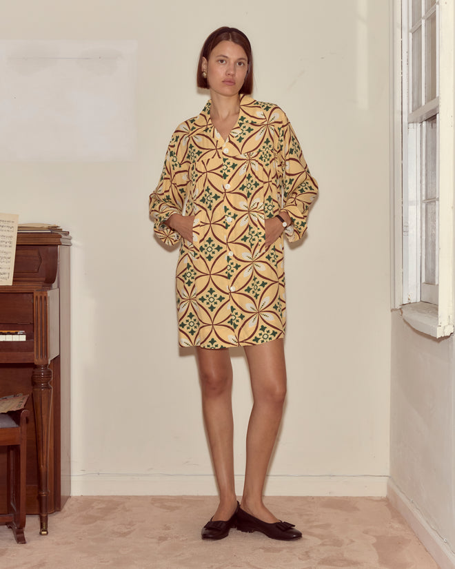A woman wearing the Greer Quincy Dress, featuring a 1960s-inspired geometric pattern, stands indoors by a window and piano with her hands in her pockets, looking at the camera.