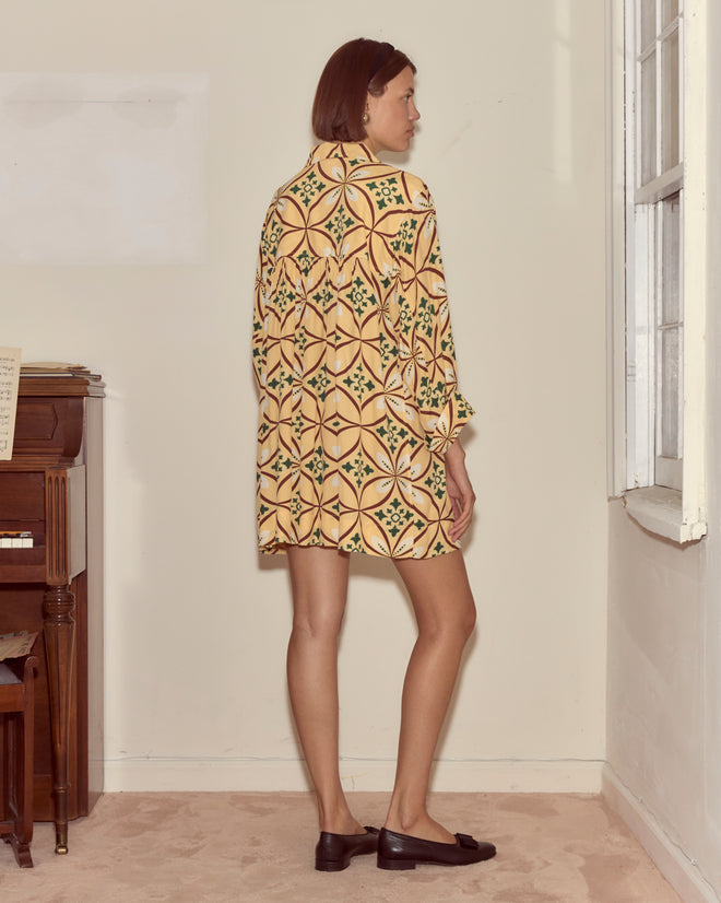 A woman in the Greer Quincy Dress, featuring a geometric pattern, stands sideways by a window in a room with a piano and beige carpet.
