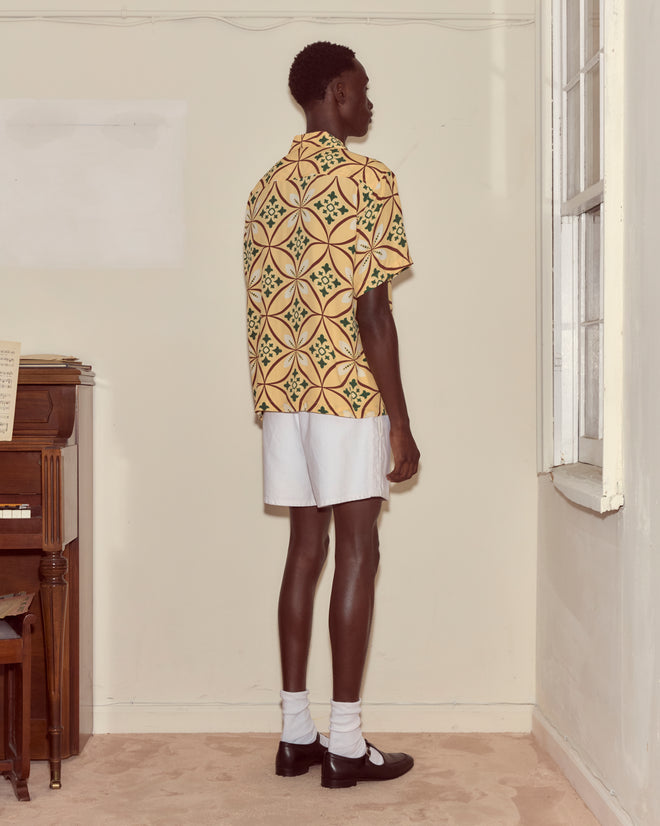 Wearing the Greer Short Sleeve Shirt with a geometric pattern and white shorts, a person stands facing a corner in a softly lit room.