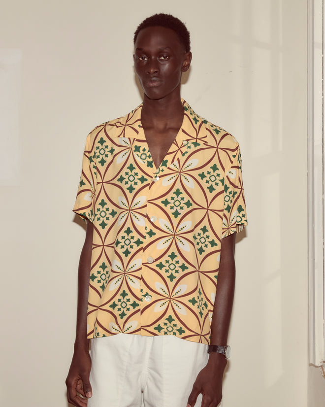 A person wearing the Greer Short Sleeve Shirt with a geometric 1960s pattern and white pants stands against a light-colored wall.
