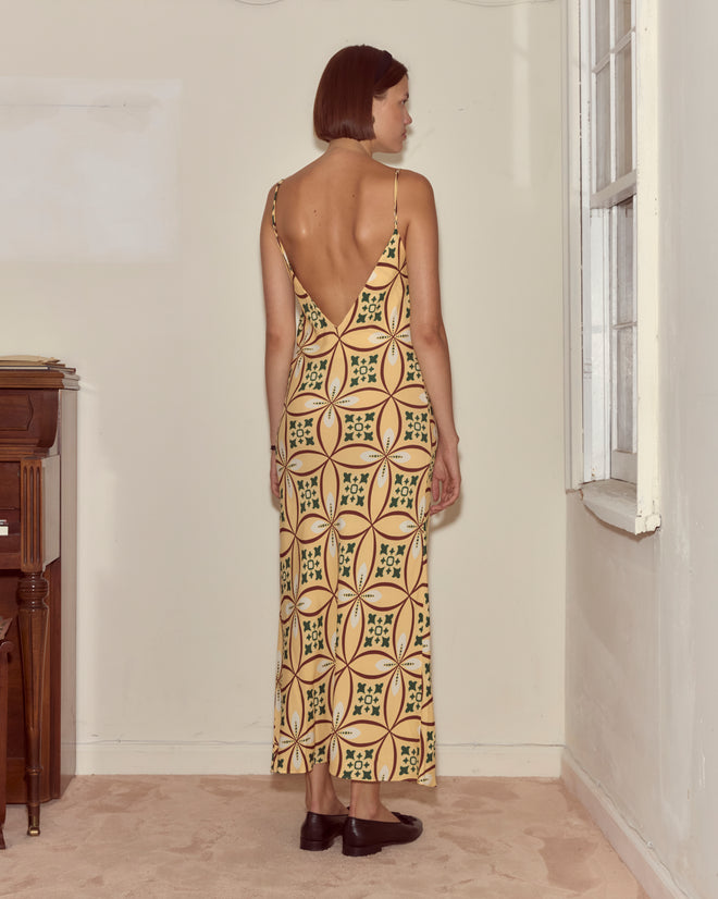 A woman with short hair stands by a window in a minimally furnished room, wearing the Greer Slip Dress with a geometric pattern.