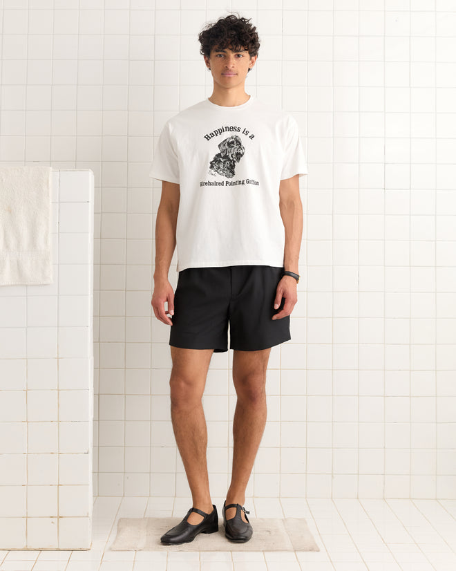 A man wearing the Happiness Tee and black shorts stands in a white-tiled room, facing the camera.