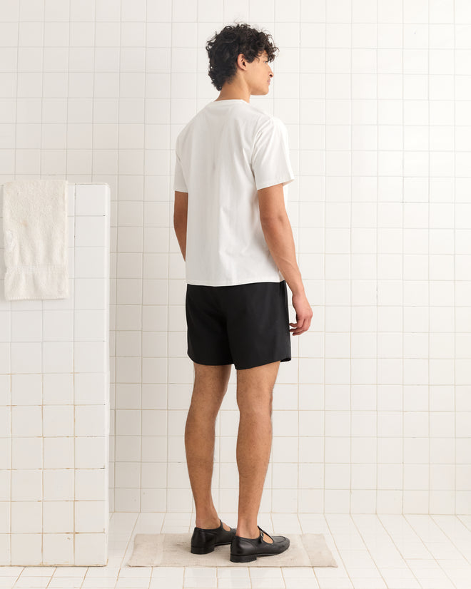 A man wearing the Happiness Tee, a 100% cotton white t-shirt, and black shorts stands on a bath mat in a tiled bathroom, facing away.