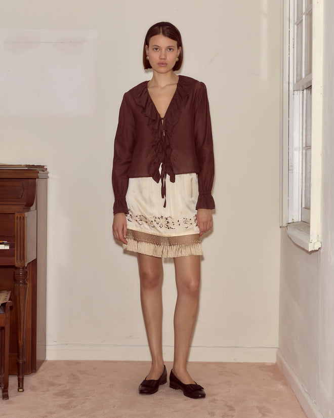 A woman in the Heartwood Flounce Blouse and a cream skirt stands indoors beside a wall and window, next to a piano.