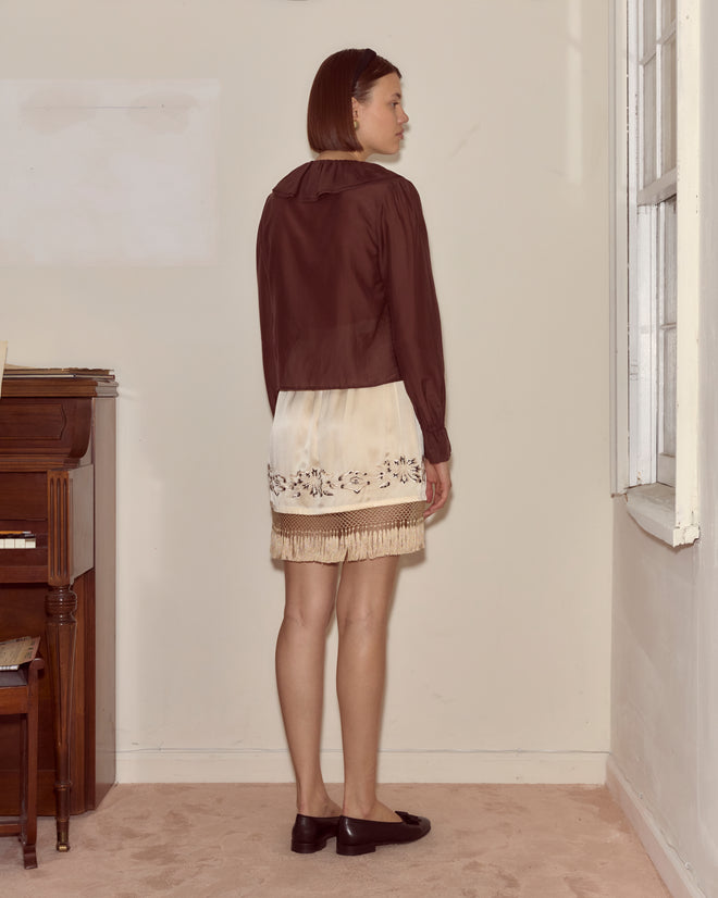 A woman with a brown bob stands sideways in a room, wearing the Heartwood Flounce Blouse in brown, paired with a cream skirt and black shoes.