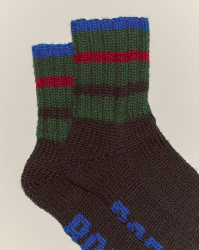 The Herrington Stripe Socks - Green feature two hand-knit socks with green, blue, and red striped cuffs and blue text on the soles.