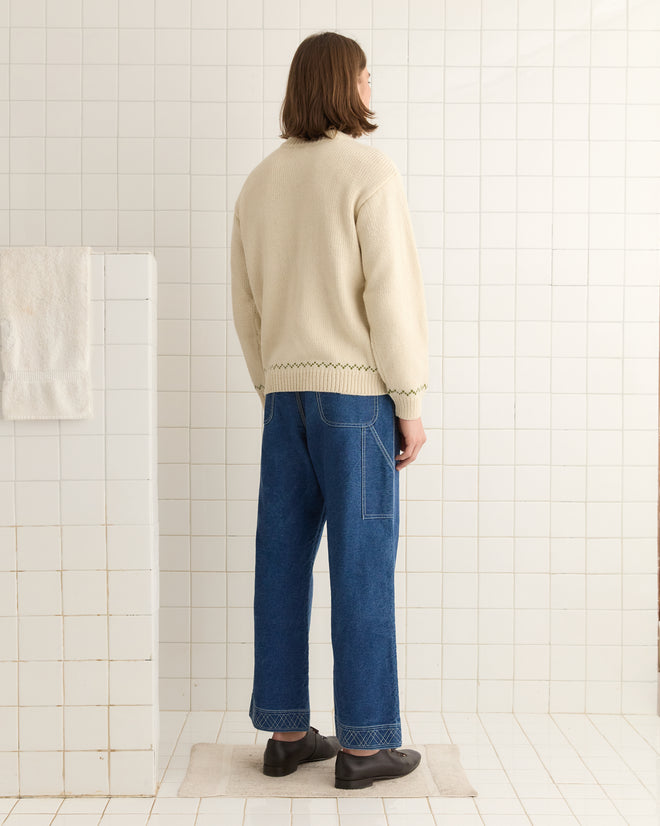 In a tiled room, a person is facing away, wrapped in the cozy Homestead Sampler Sweater, paired with blue jeans and black shoes.