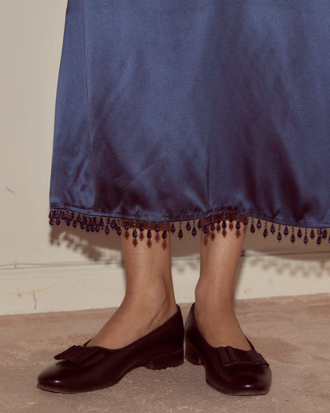 A person stands on a beige carpet wearing the Hypnos Dress, a blue deadstock satin piece with beaded trim, paired with black loafers.