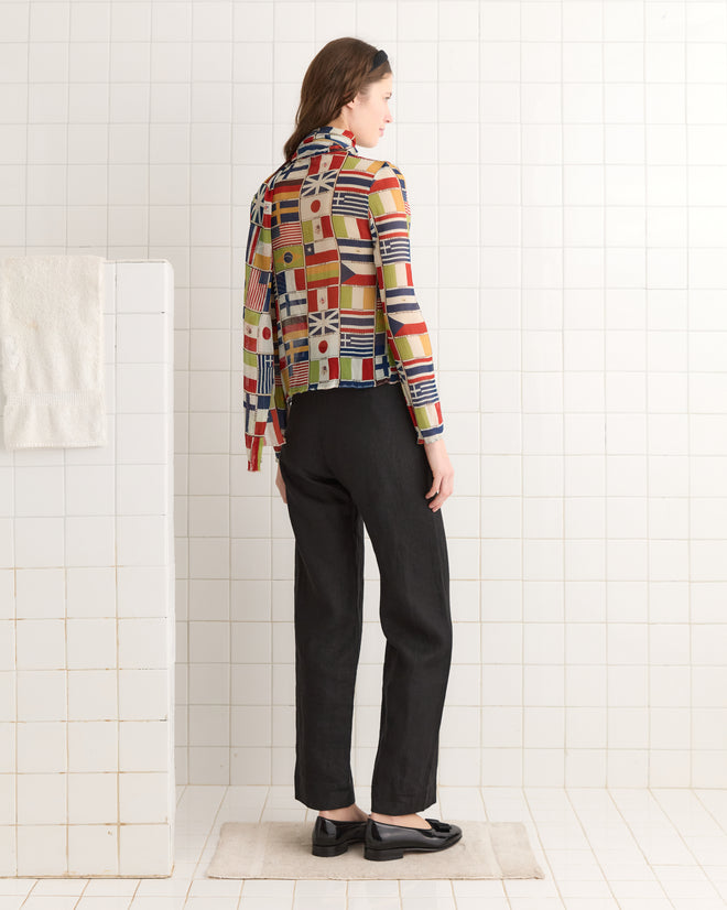 A person in an International Blouse with a colorful flag print and black pants stands in a white tiled room.