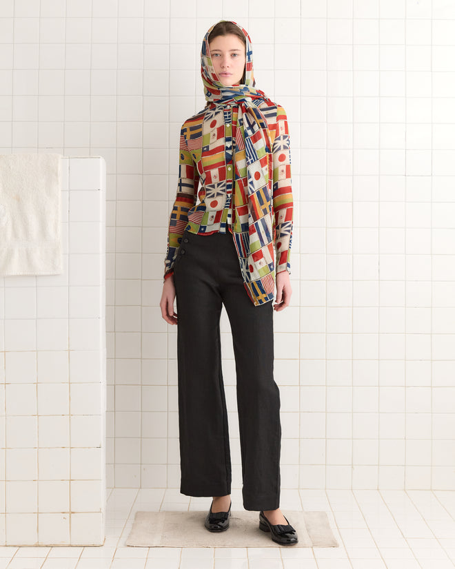 Against a tiled wall, someone stands gracefully in an International Blouse—an eye-catching flag-print top paired with a scarf tie—and classic black pants.