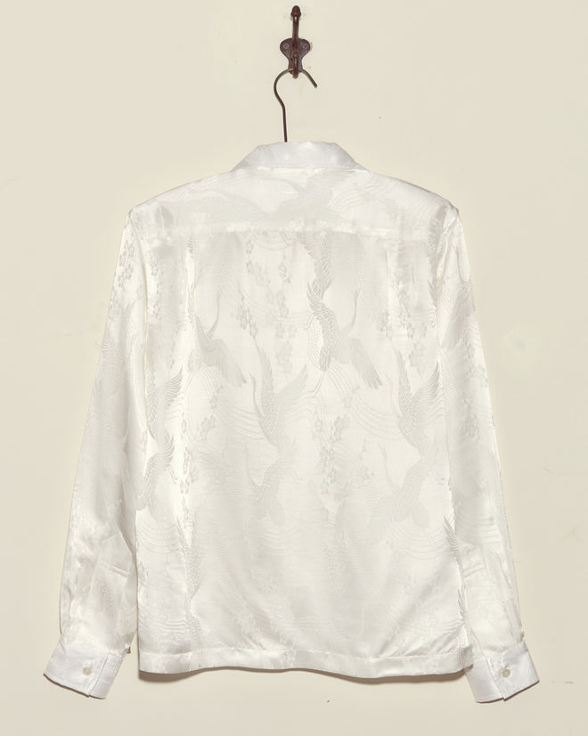 The Ivory Cloud Long Sleeve Shirt, crafted from white deadstock jacquard with a subtle crane pattern, hangs on a hook against a plain wall.
